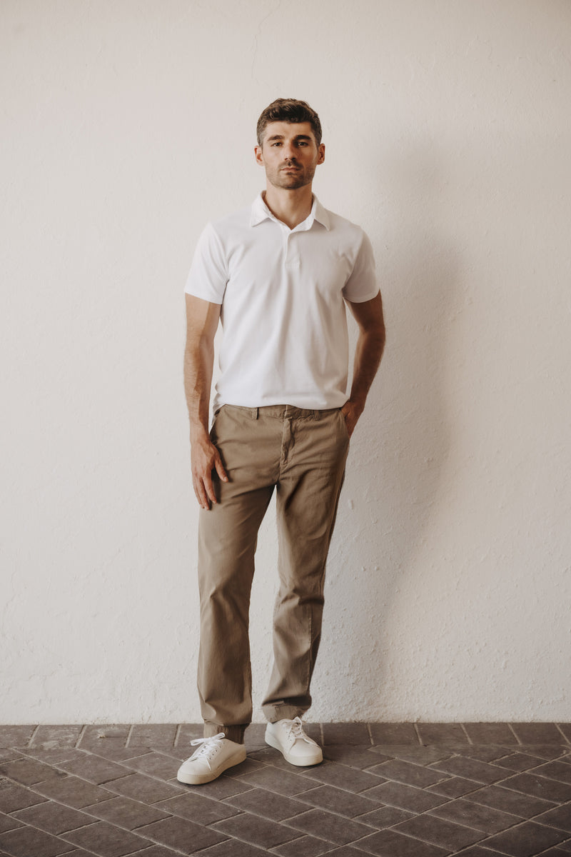 A man wearing a SKU white short-sleeve polo shirt, SKU standard chino khaki trousers, and white sneakers stands against a textured off-white wall.