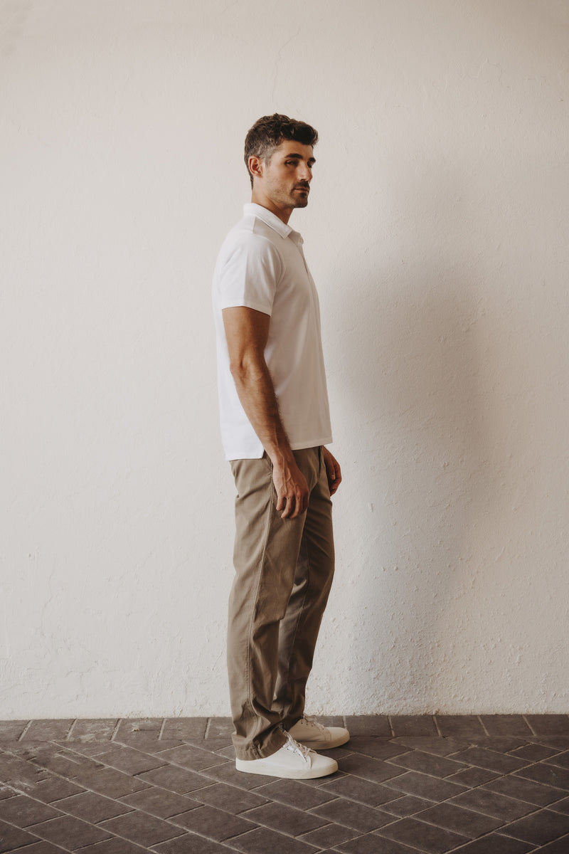 A man standing in profile wearing a SKU white short-sleeve polo shirt, SKU standard chino khaki trousers side view, and white sneakers stands against a textured off-white wall.