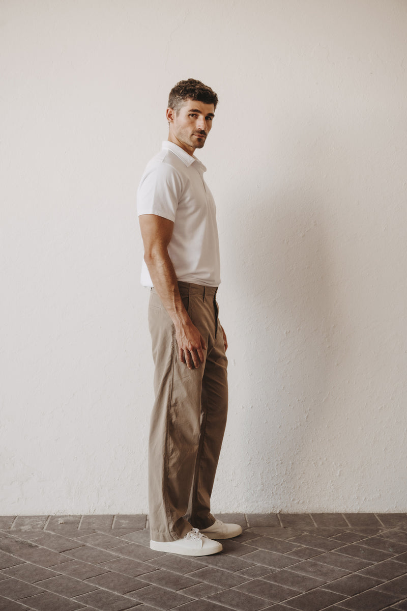A man wearing a white short-sleeve polo shirt and Save Khaki United Weekend Chino in khaki, standing in profile against a textured off-white wall on a dark tiled floor. This image show the straight leg fit on model in side view.