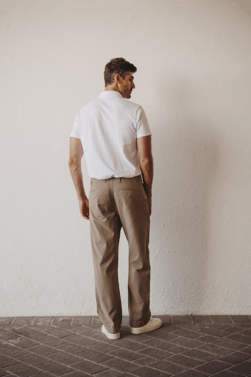 A man stands with his back to the camera wearing a white short-sleeve polo shirt and beige trousers, standing on a dark tiled floor against a textured off-white wall. This image shows the Save Khaki United Weekend Chino in back view.  It is a straight leg with loose hip and thigh and straight to the leg opening.