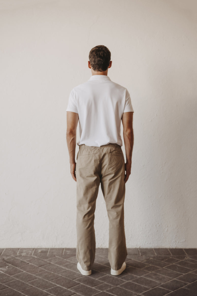A man viewed from behind wearing a white short-sleeve polo shirt and SKU easy chino in khaki, standing against a plain off-white wall on a dark tiled floor.