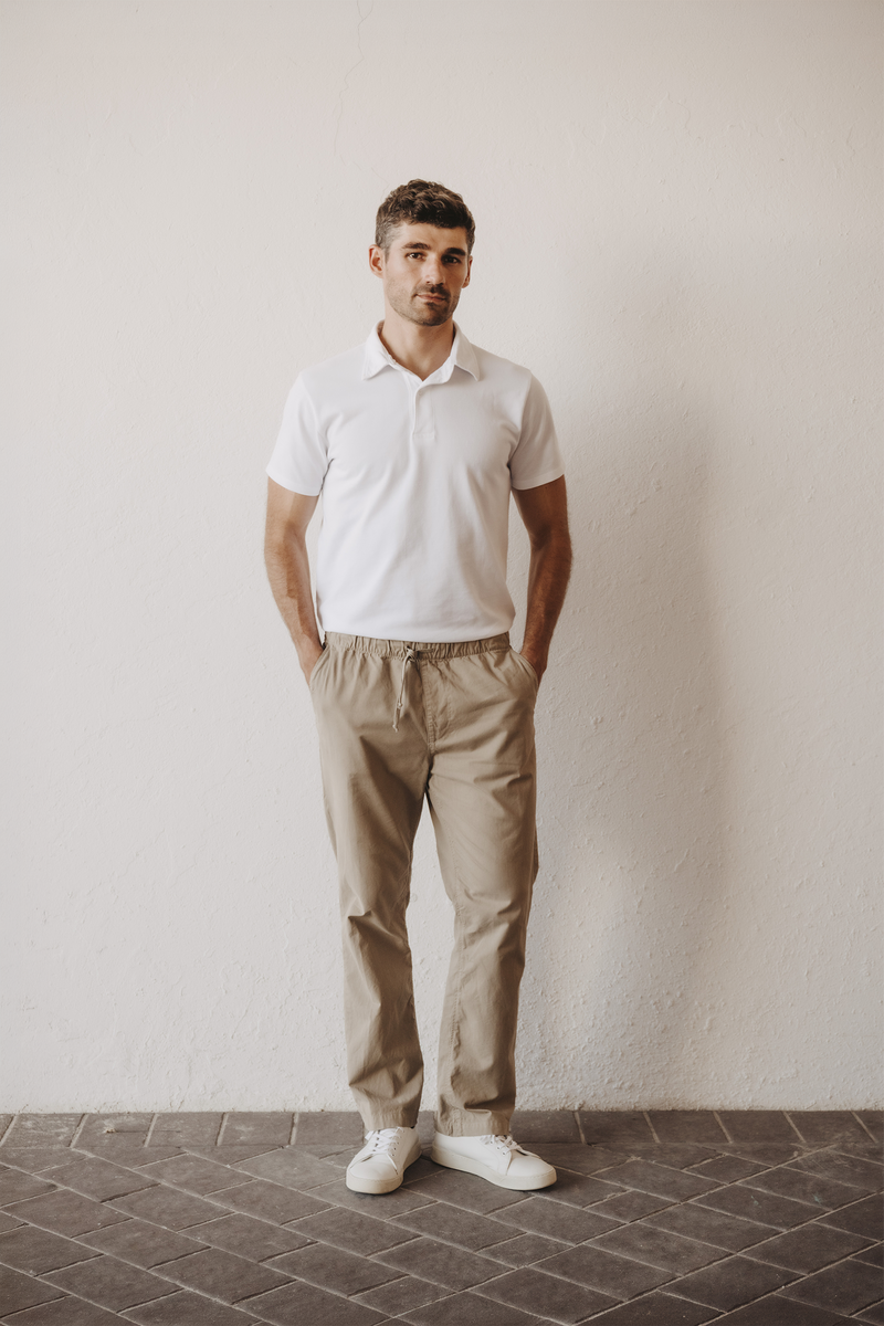 A man wearing a white short-sleeve polo shirt, beige drawstring trousers, and white sneakers, standing against a textured off-white wall with a dark tiled floor.  Model wearing Save Khaki United Easy Chino with elastic waistband that is comfortable on the waist, hip, thigh, and taper to the leg for a slimming fit. 