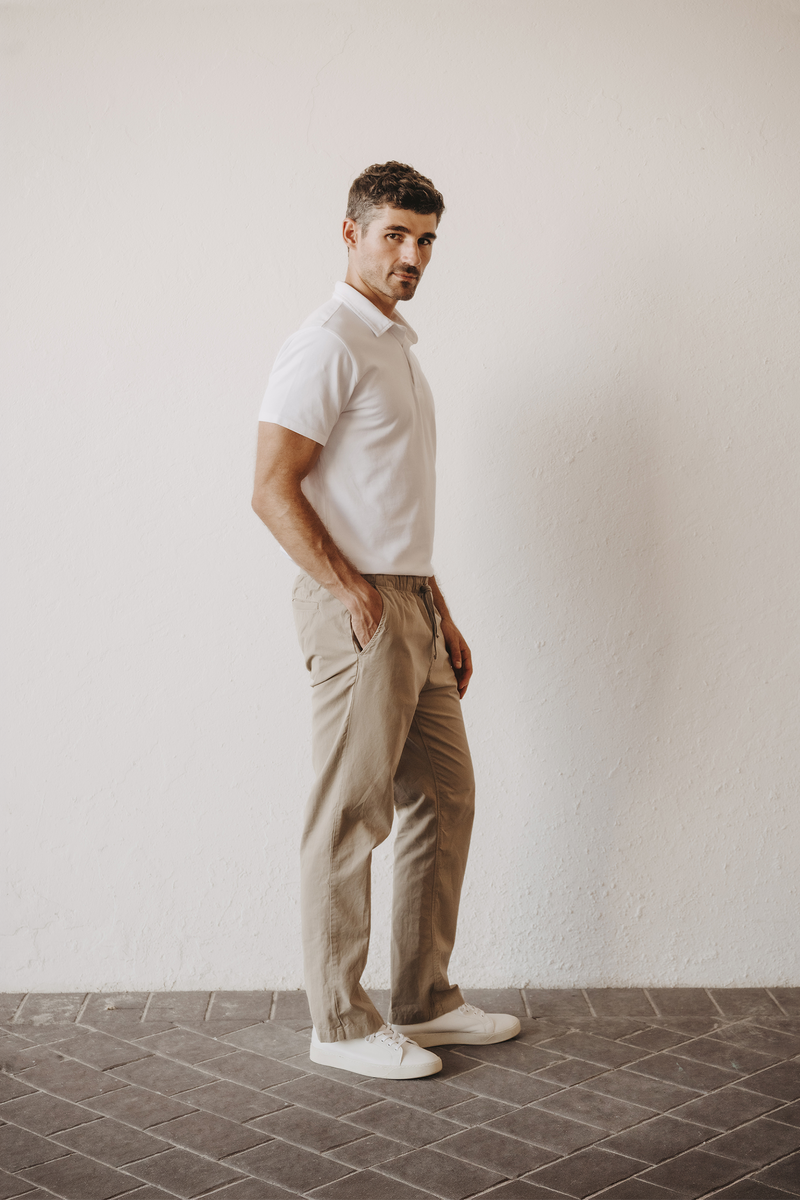 A man in a white short-sleeve polo, Save Khaki United Easy Chino in khaki and white sneakers, shown in profile. Elastic waistband and a tapered leg.