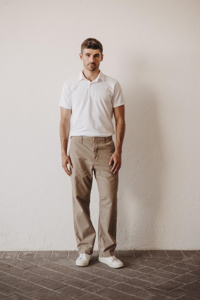 A man stands against a textured off-white wall wearing a white short-sleeve polo shirt, Save Khaki United Weekend Chino in khaki, and white sneakers.  This is a straight leg chino, mid rise.  Trouser sits comfortable on the waist with loose thigh and straight leg opening.