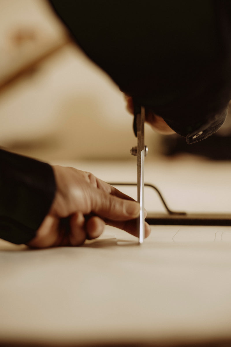 A close-up shot of a person's hands using a metal divider or compass tool on a light surface, with a dark sleeve and blurred warm-toned background. Save Khaki United makes all its chinos in Los Angeles with imported fabric.