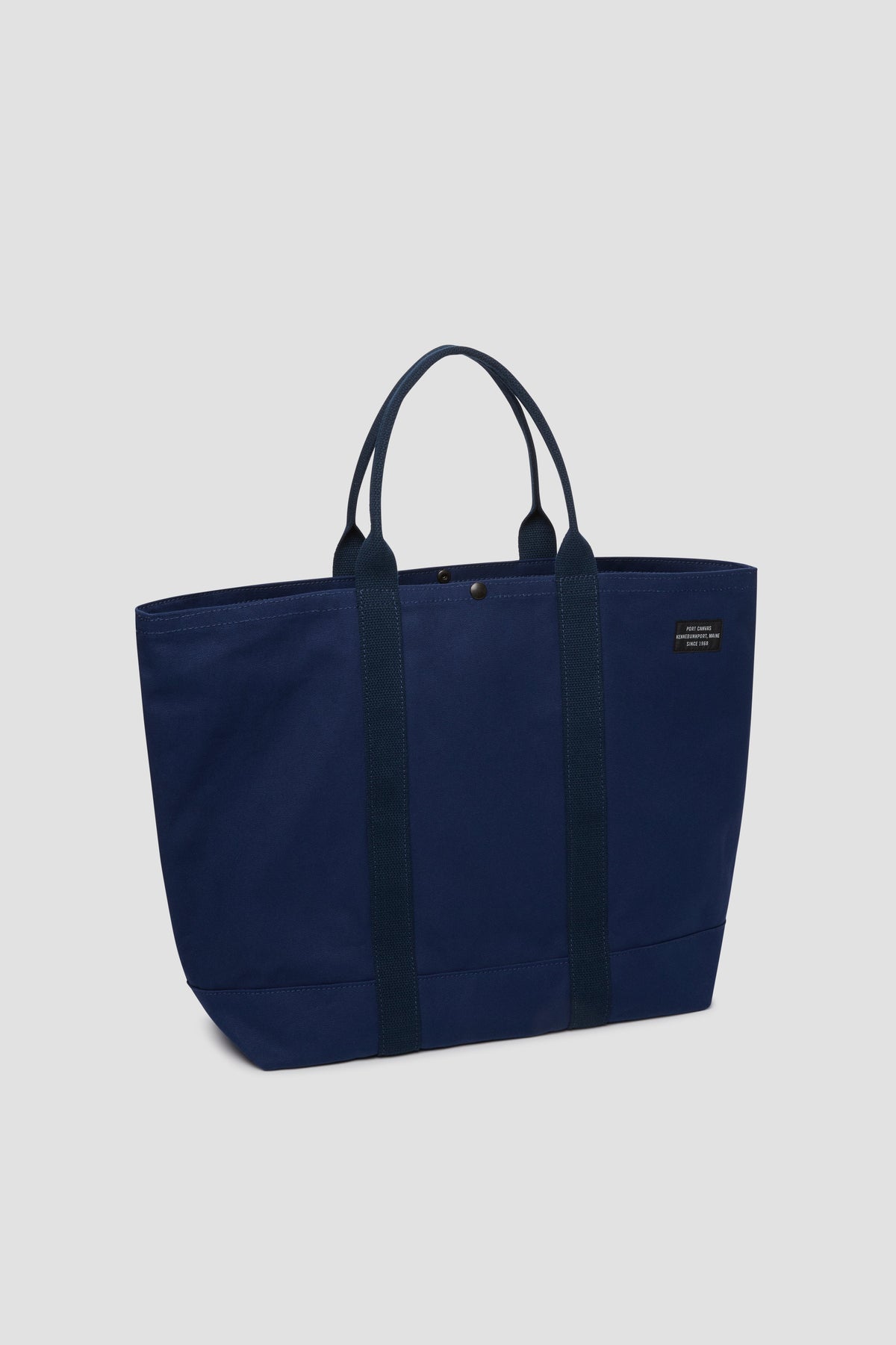 Navy canvas tote bag with top button, short handles, and front patch logo.