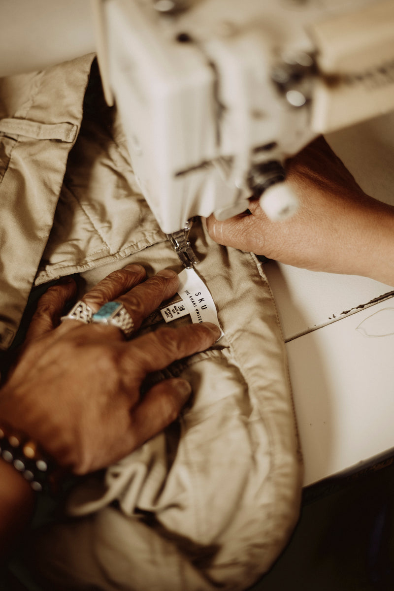 A close-up shot of a person's hands guiding a beige quilted garment under a sewing machine, with a small sewn label visible. This image shows the Save Khaki United label on the interior of the Chino.